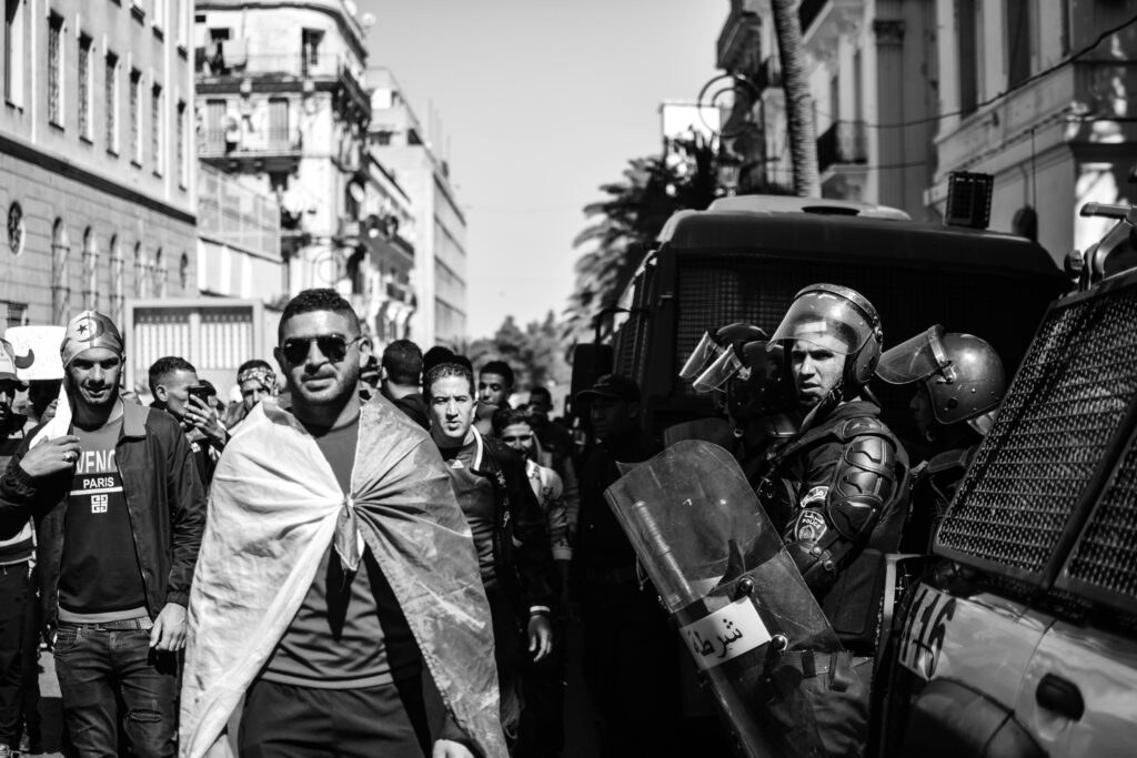 Black and white scene of protesters and police in an Algerian street, highlighting tension.