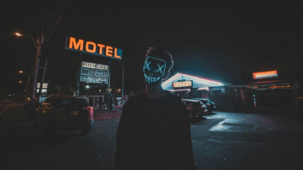 A spooky urban scene at night with a person wearing a glowing LED mask near a motel.