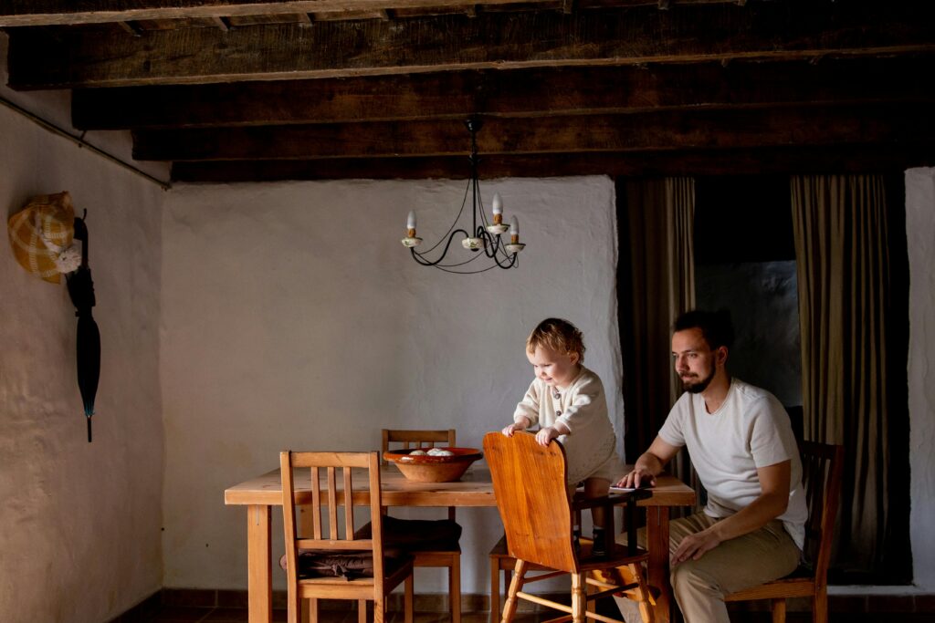 Cheerful cute little daughter spending time together with bearded father in casual wear inside aged room near wooden dinner table with wooden chairs at daytime