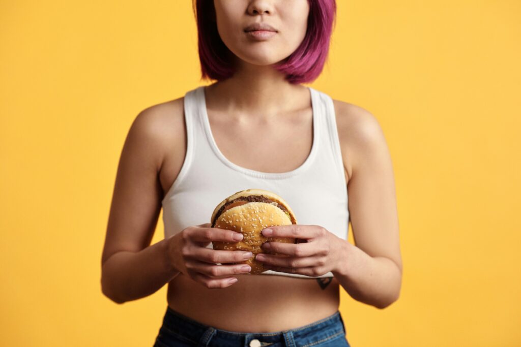 Young woman with pink hair holding a burger, set against a yellow backdrop.