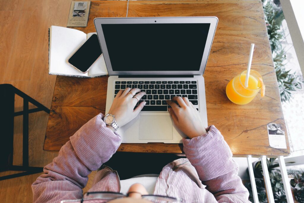 A person wearing pink sleeves works on a laptop at a wooden desk with a notebook, phone, and juice.