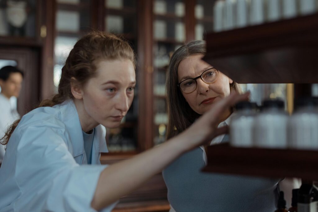 Two pharmacists wearing white coats and glasses examining medicine bottles on a pharmacy shelf.