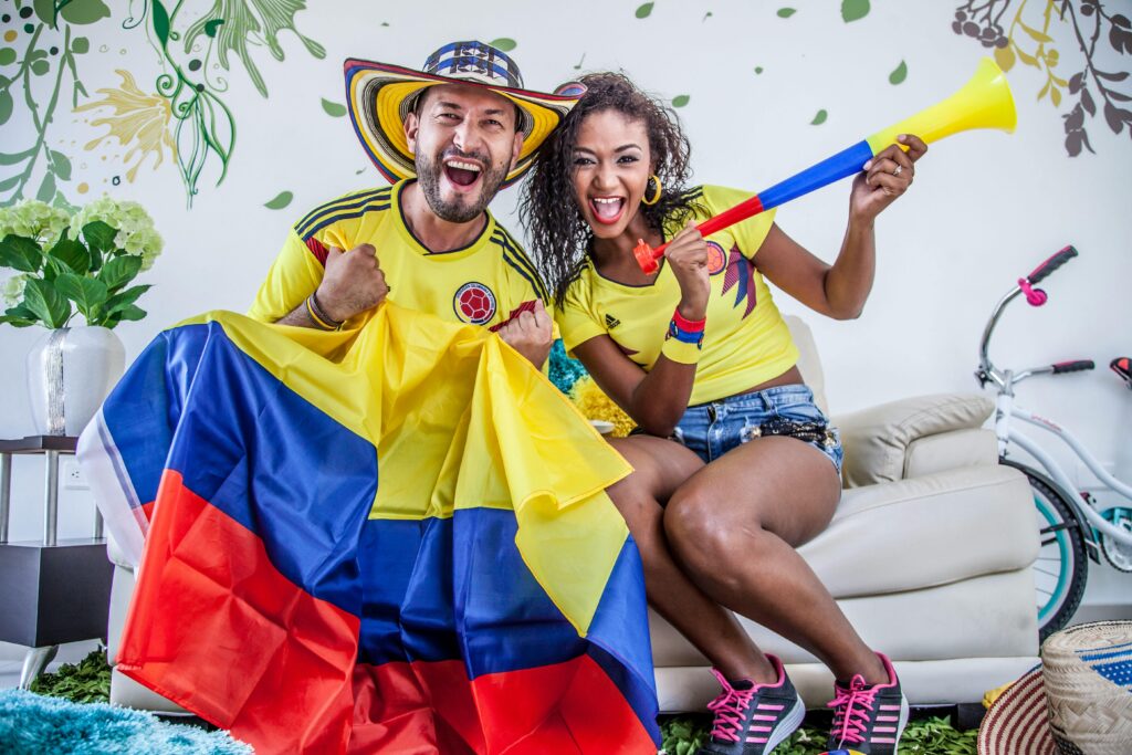 Joyful Colombian couple celebrating team victory with flags and vuvuzela indoors.