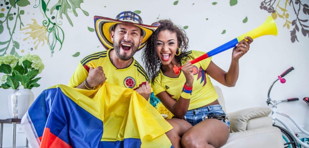 Joyful Colombian couple celebrating team victory with flags and vuvuzela indoors.