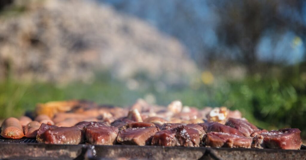 Close-up of various meats sizzling on a barbecue grill outdoors on a sunny day, showcasing a summer cooking scene.