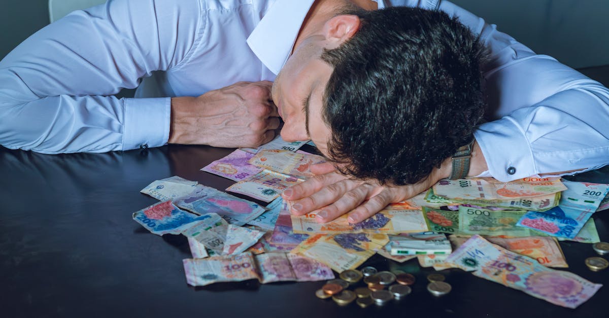 A tired businessman rests his head on a table covered with Argentine pesos and coins.
