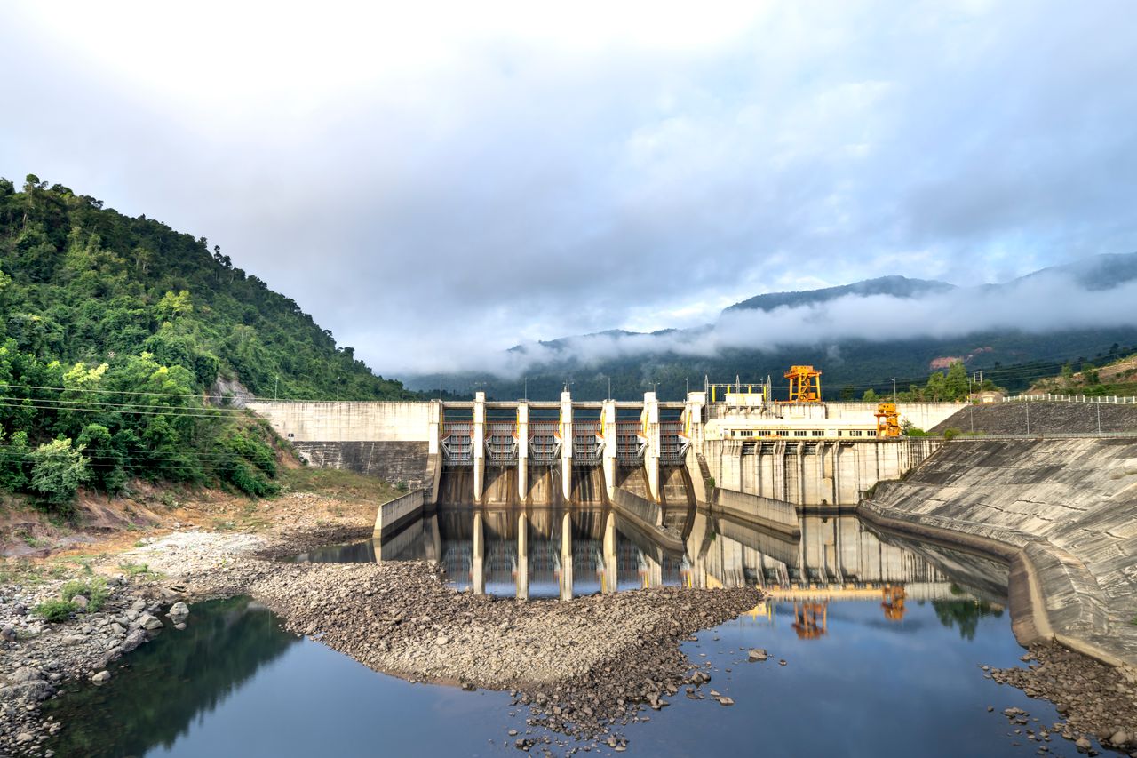 Industrial hydroelectric power plant located near river and stony coast against green mountain range covered with mist on summer day